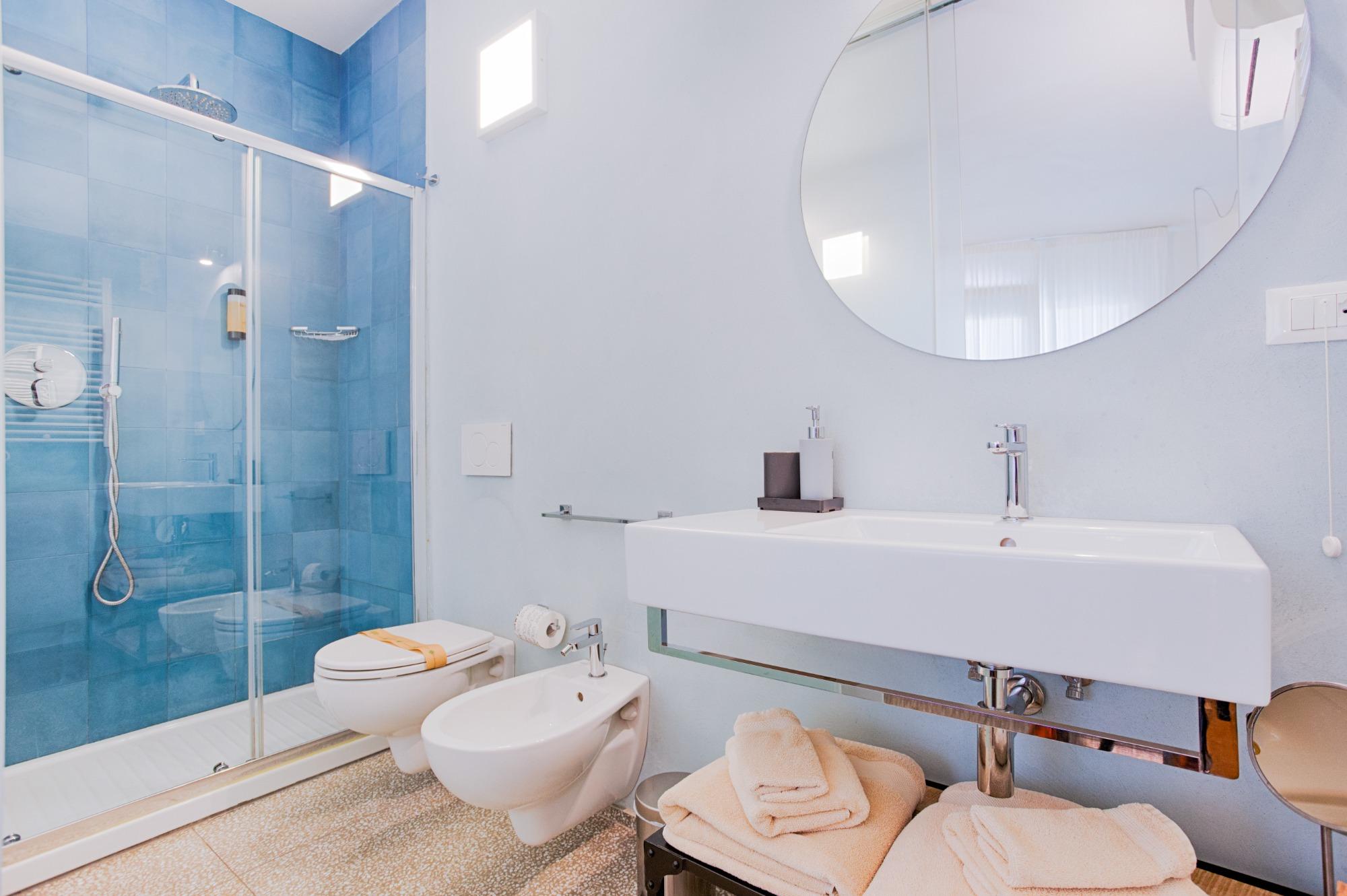 CamerBagno privato interno della Camera Azzurra con grande doccia blu, sanitari sospesi e lavandino moderno.a Sabbia  con letto matrimoniale e bagno